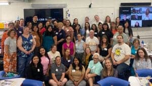 A large group of diverse educators smiling together in a classroom. Participants are seated and standing, with visible name tags, math materials on tables, and digital displays in the background showing remote attendees.