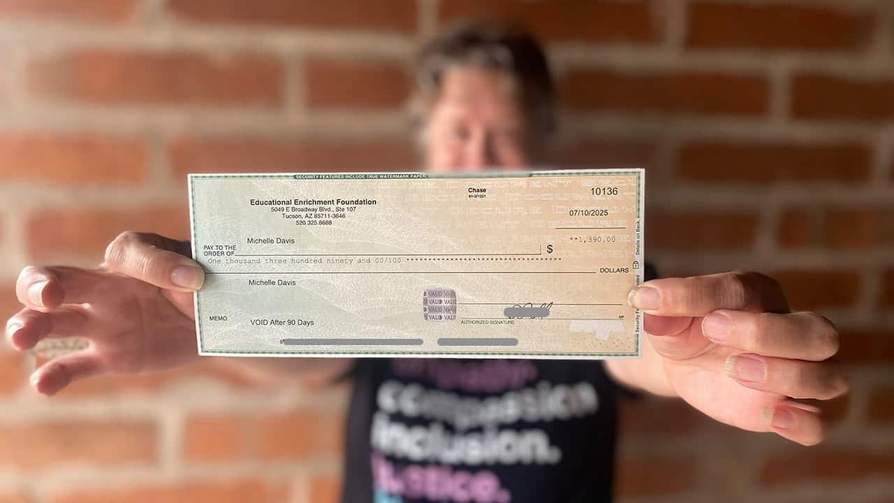 A person holds up a check from the Educational Enrichment Foundation, issued to Michelle Davis for $1,390.00, dated July 10, 2025. The background shows a blurred brick wall, while the focus is on the check in the foreground.
