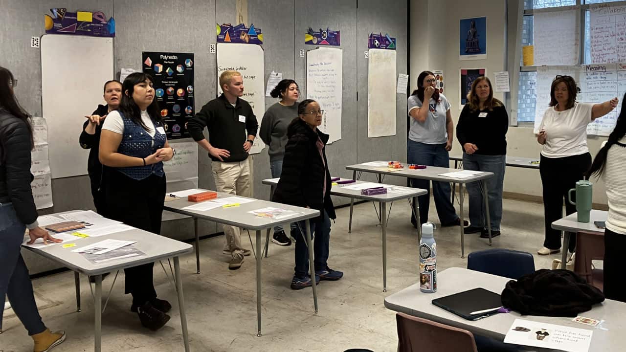 Adults stand and interact around tables in a classroom setting, with papers, name tags, and educational posters visible on the walls.